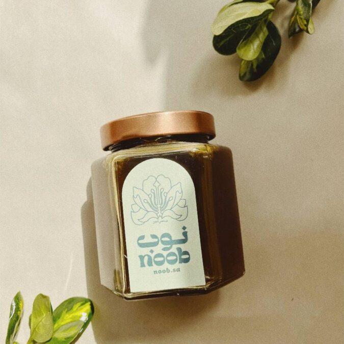 مرطبان زجاجي سداسي الشكل يحتوي على عسل سدر، مع غطاء نحاسي لامع، موضوع على سطح بيج اللون مع أوراق خضراء صغيرة في الخلفية. شعار "نوب" واضح على ملصق أبيض على المرطبان، يحيط به تصميم نباتي بسيط.